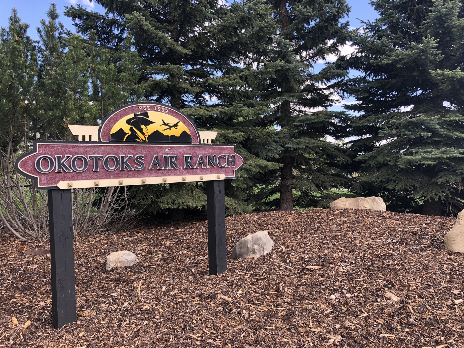 Okotoks Air Ranch entrance sign among mature spruce trees, established 1999